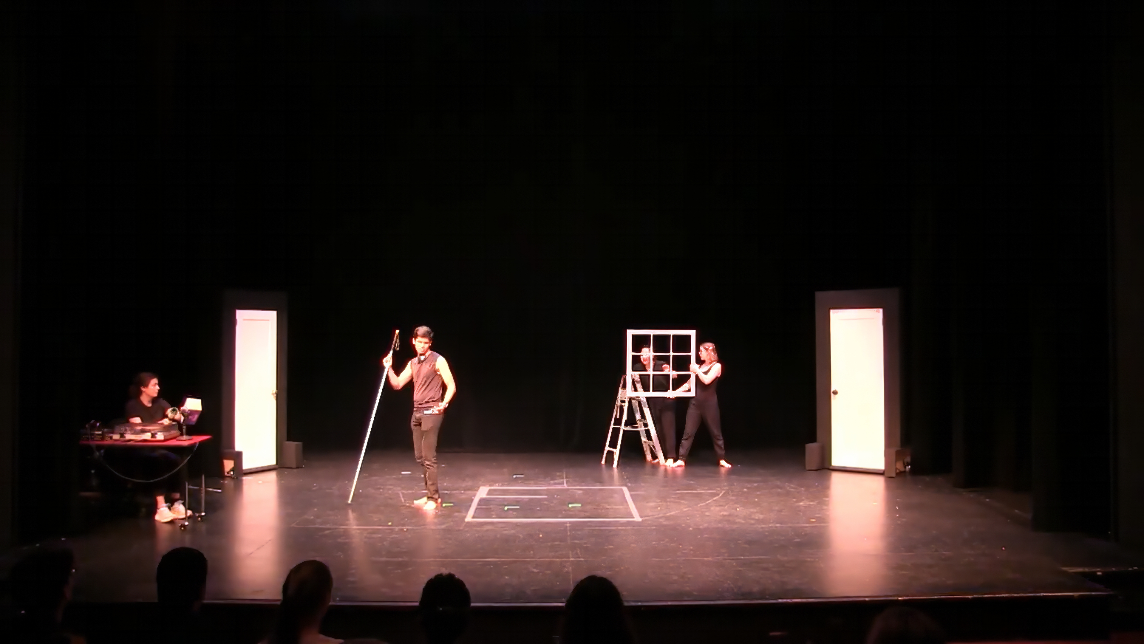 Scene from a darkened theater showing Bhavya, center stage and dressed in a sleeveless black shirt, holding his long white cane in one hand and other hand in his pocket; on the left, a woman at a soundboard table; and on the right, two performers holding a stepladder and a square window frame